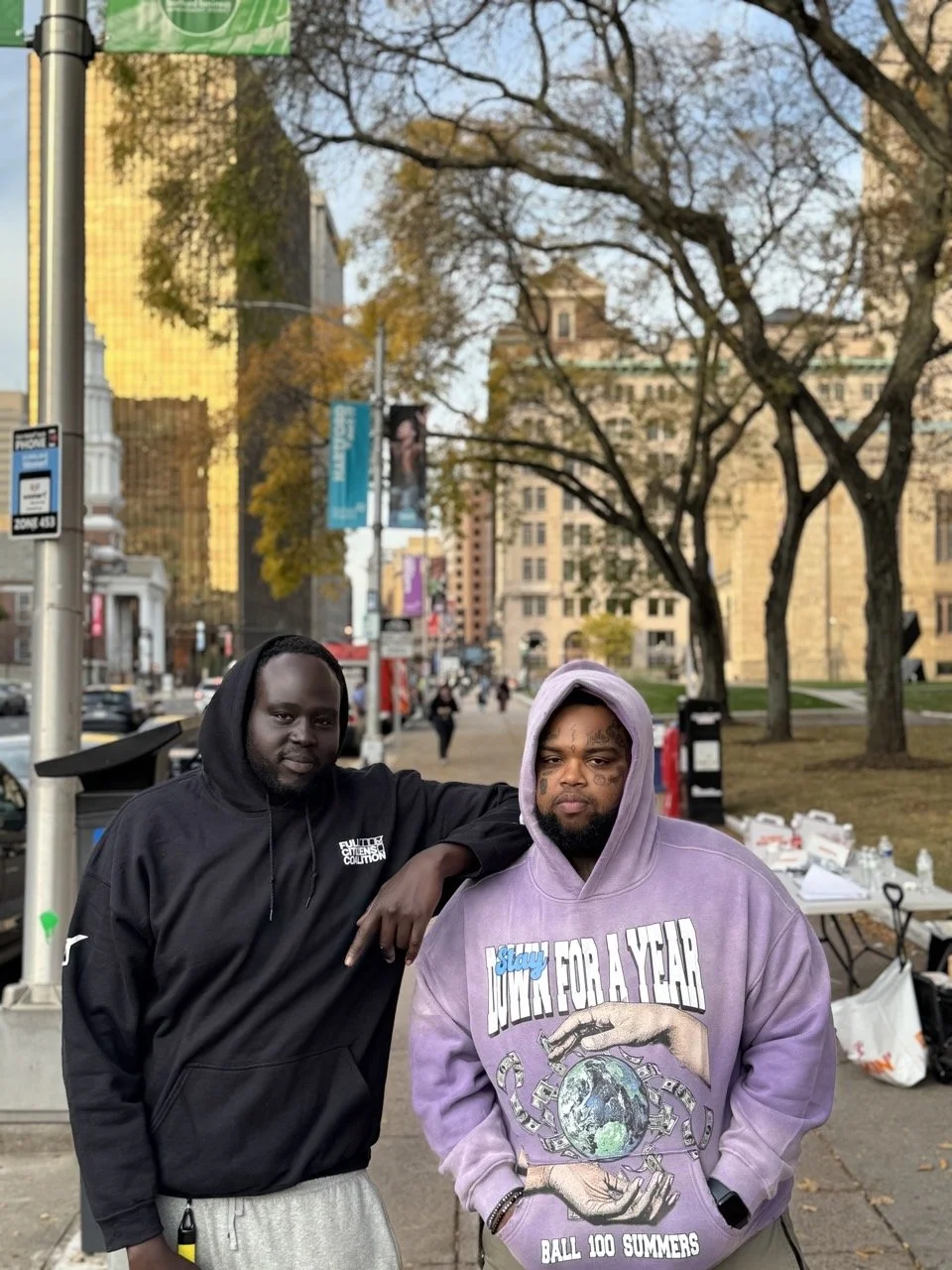 Two men, one in an FCC hoodie, on a downtown sidewalk beside an outreach table