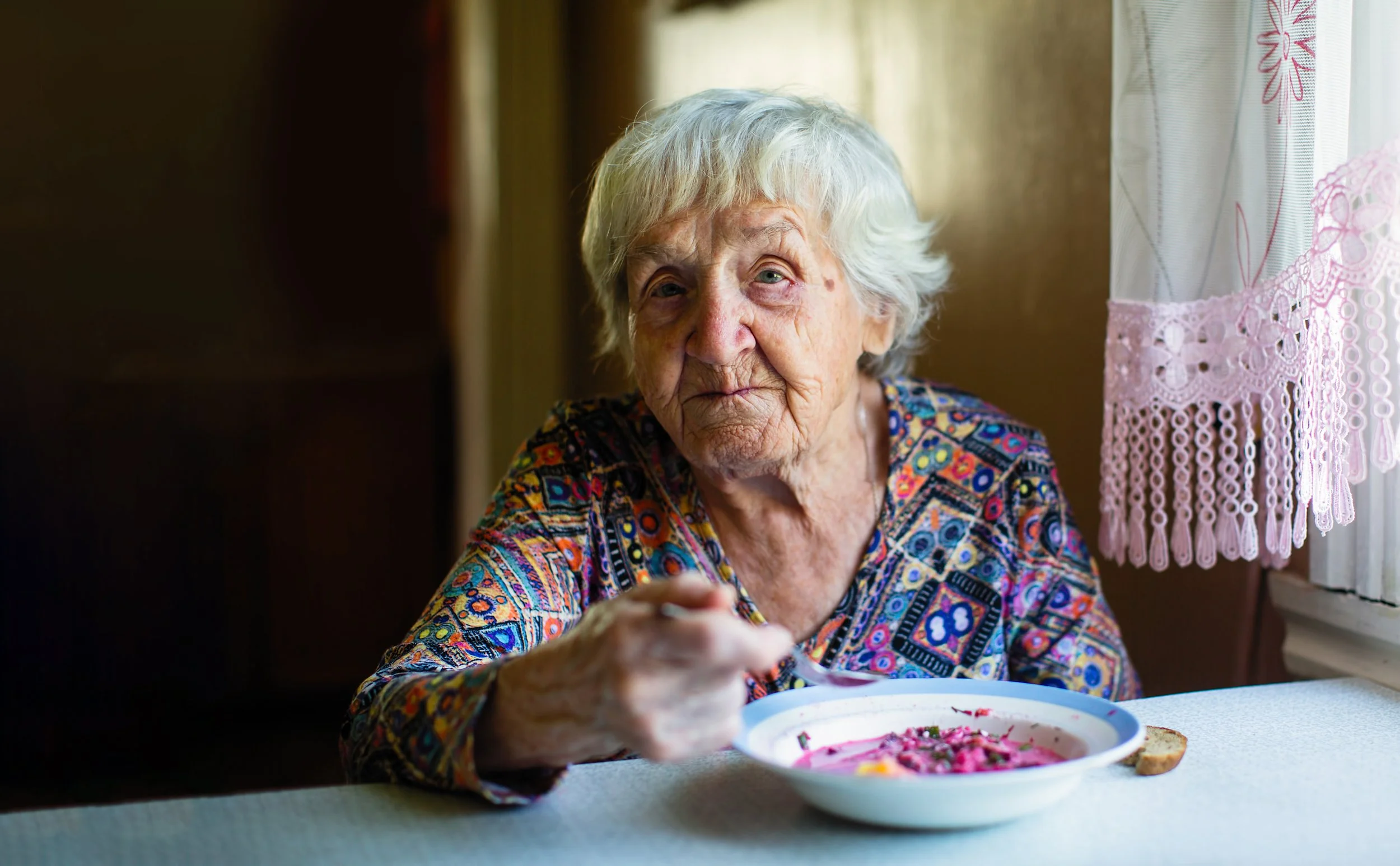 An elderly woman seated at a table eating a bowl of soup
