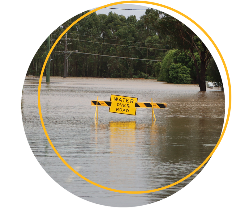 A yellow Water Over Road sign standing in floodwater that covers a tree-lined road