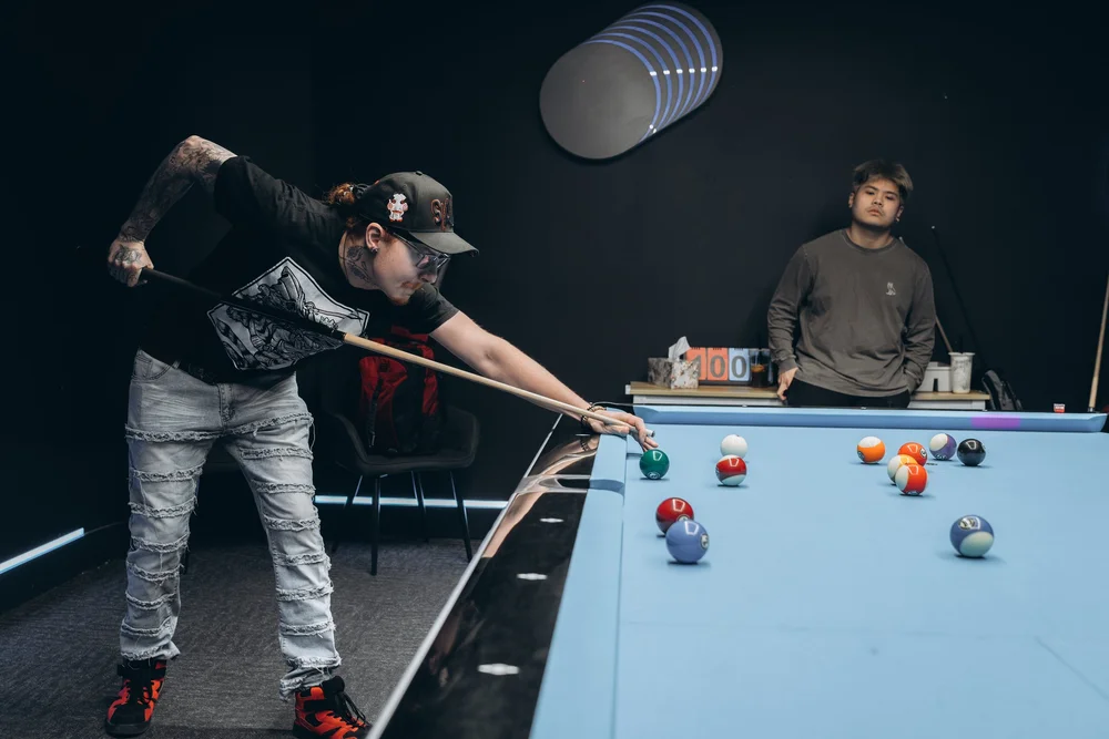 Player shooting in a game of 8-ball with Aramith Tournament balls on a Rasson 9ft table with Simonis 860HR blue cloth while a friend watches