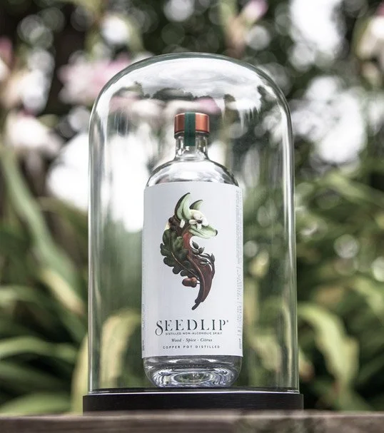 Seedlip nonalcoholic spirit bottle displayed beneath a glass cloche in a garden setting.
