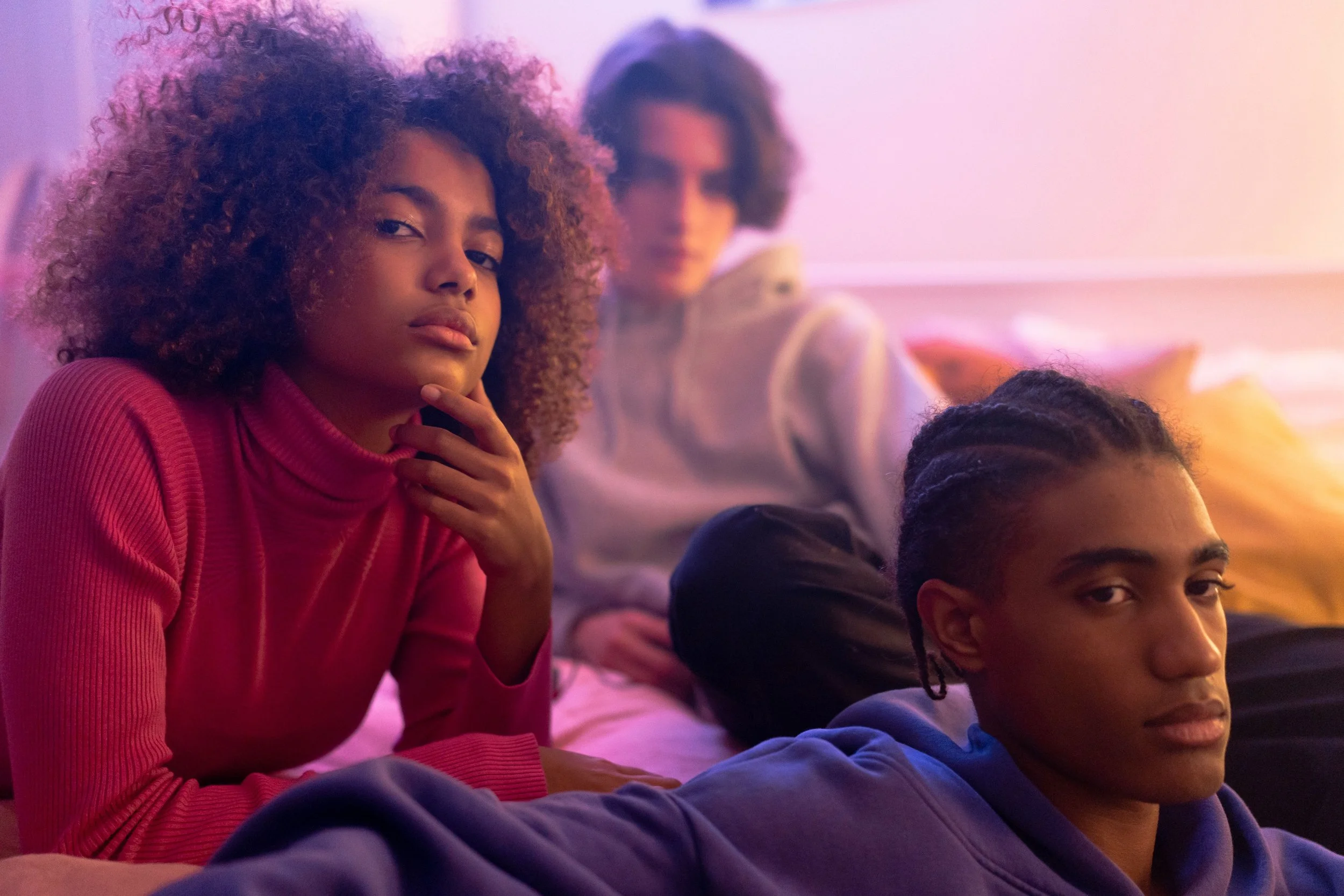 Three teenagers relaxing together in a bedroom washed in pink and purple light, each looking thoughtful.