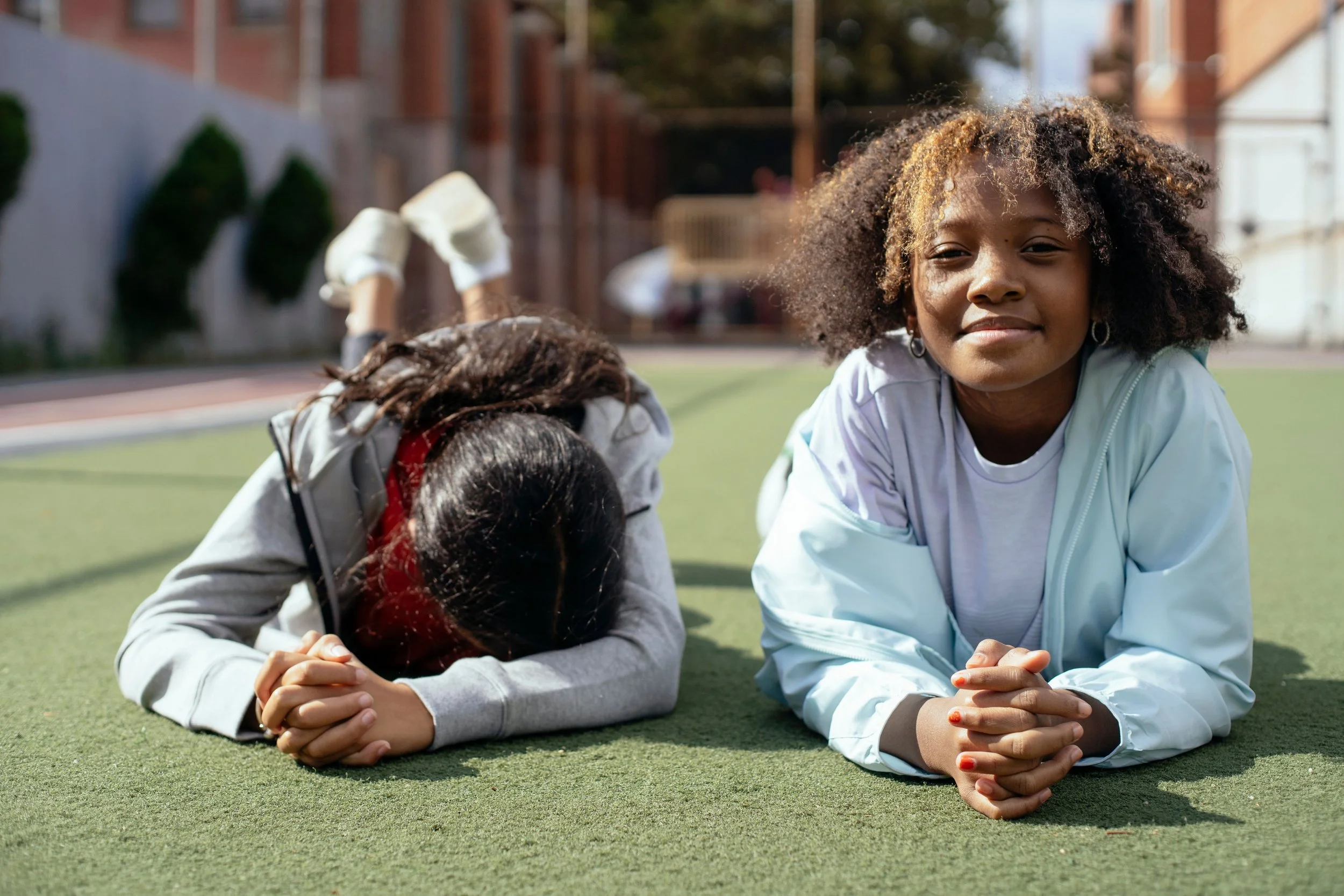Two teenage girls lying side by side on a sunlit turf field, resting on their arms and smiling.