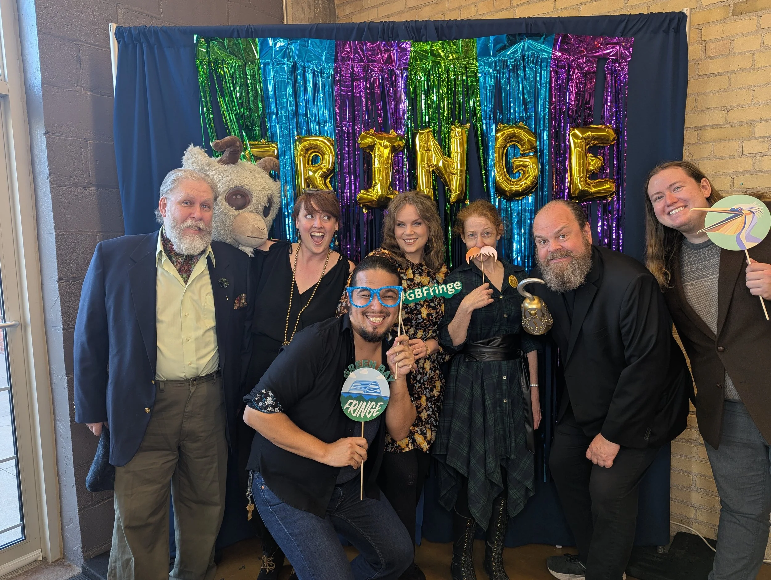 The GB Fringe board of directors hold signs in front of a colorful backdrop where Fringe is spelled with balloons