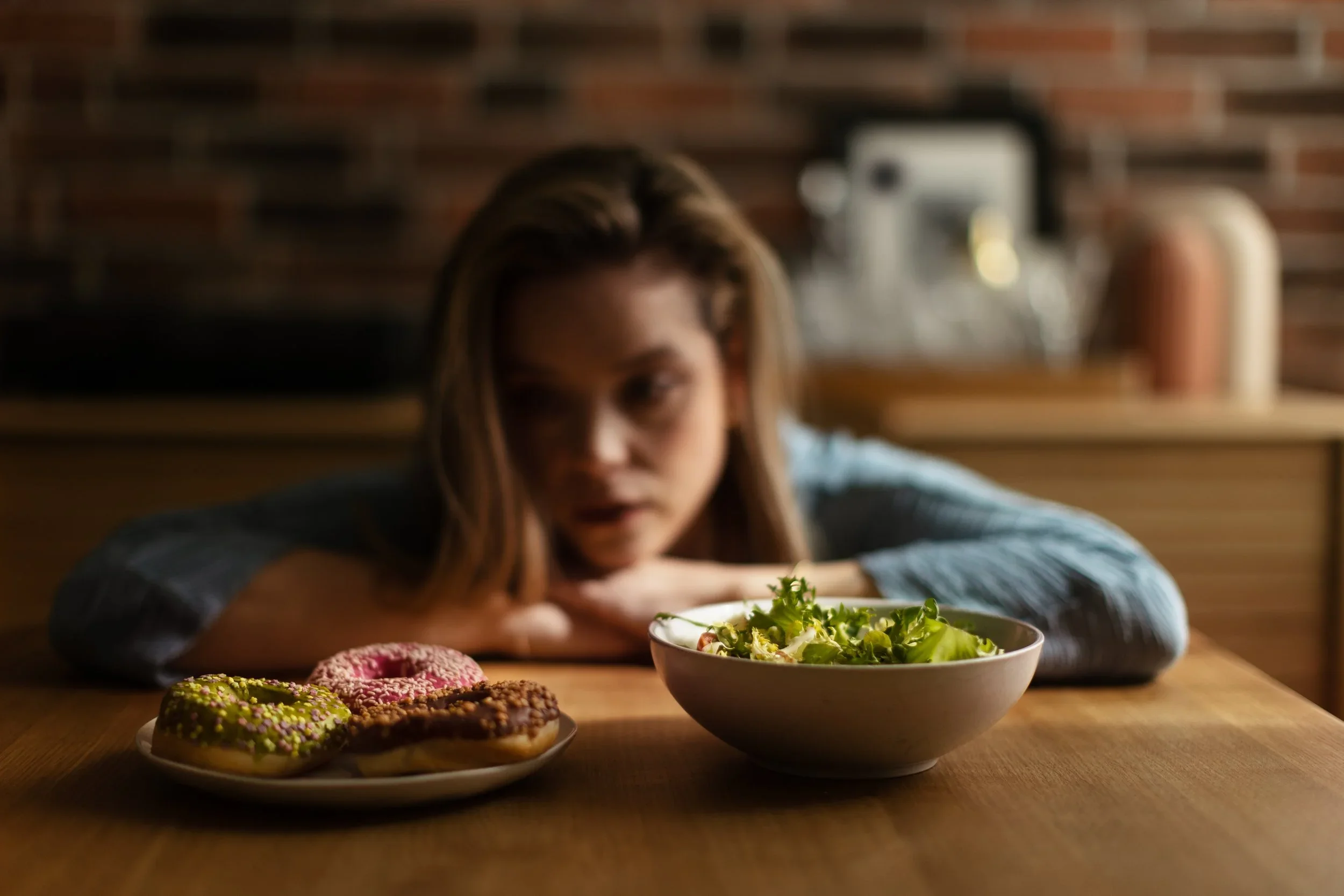 Young woman weighing food choices while seated between a bowl of salad and pastries