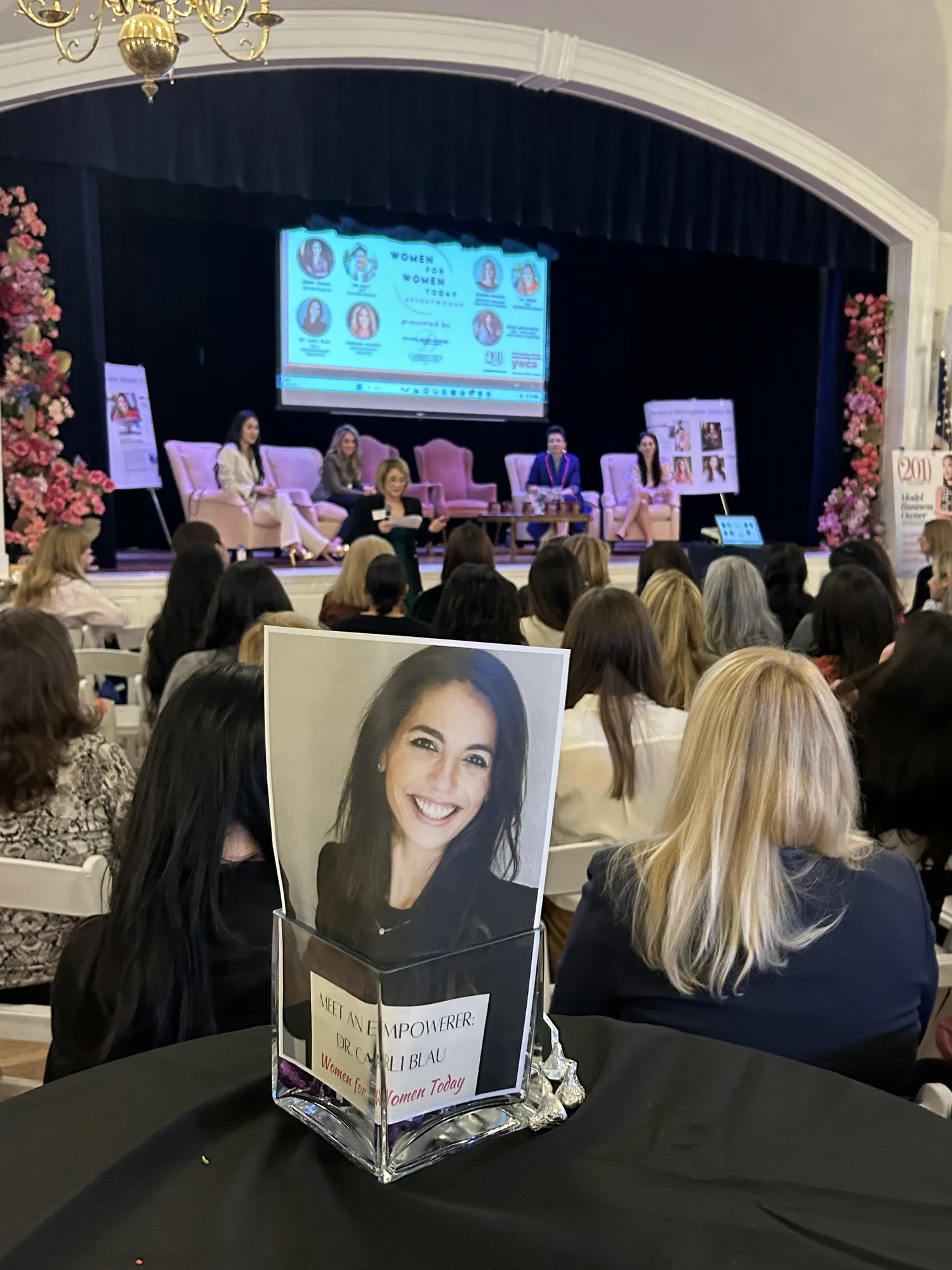 Dr. Carli Blau's table card at the Women for Women Today panel, with panelists seated on stage behind it