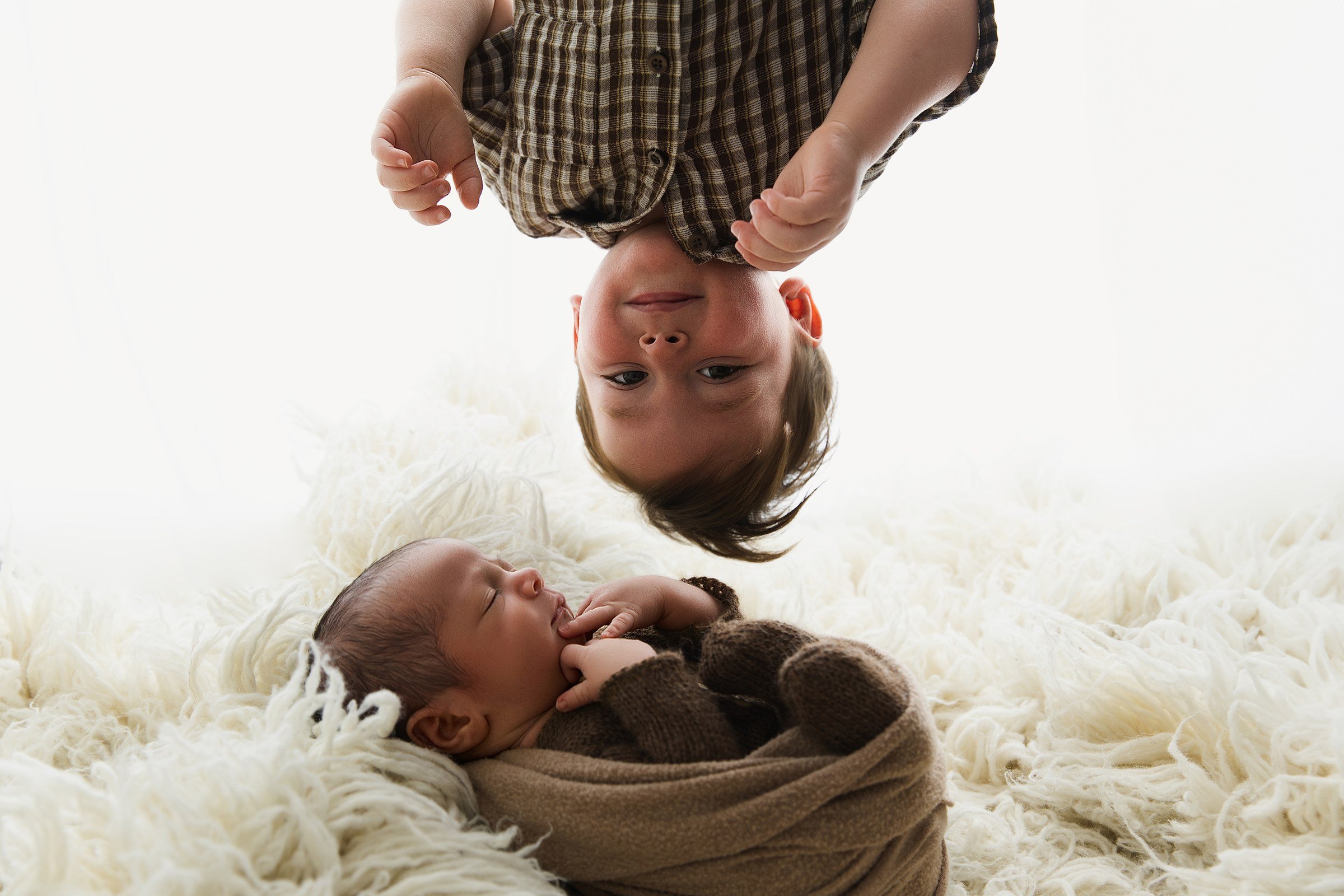 An upside-down big brother leaning into the frame beside his sleeping newborn sibling.