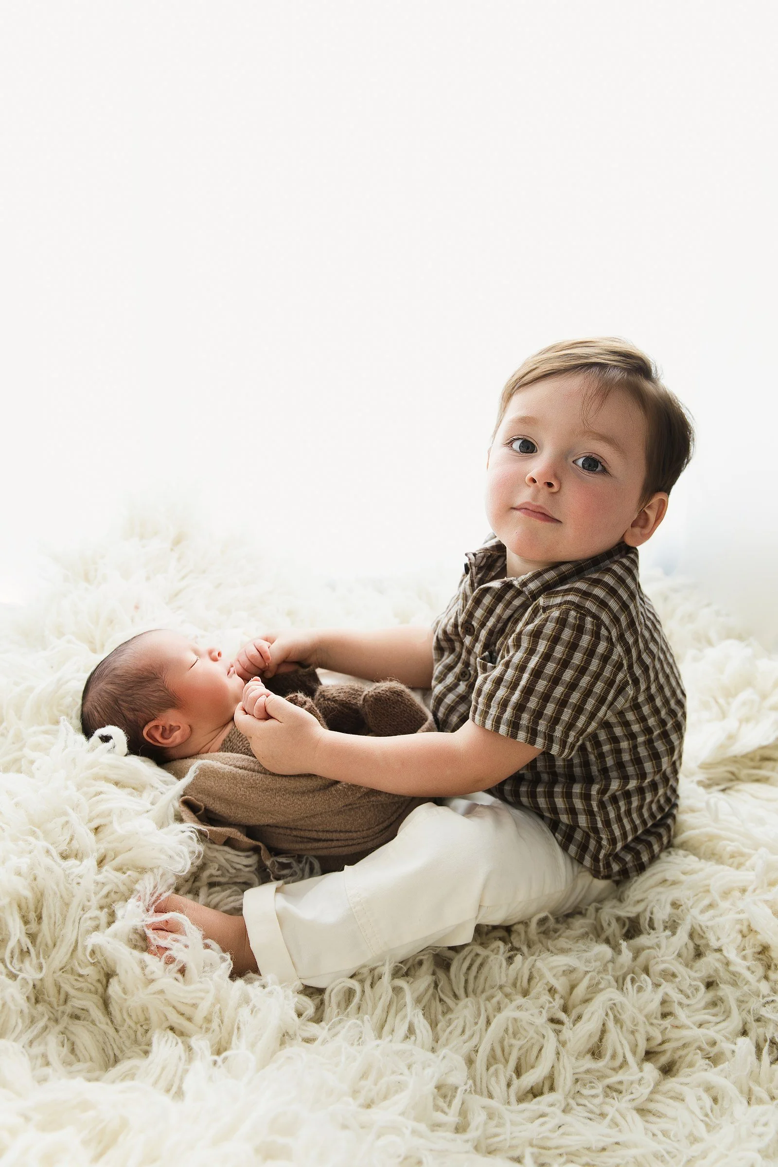 Big brother sitting close to his newborn sibling, who is wrapped in a soft brown fabric.