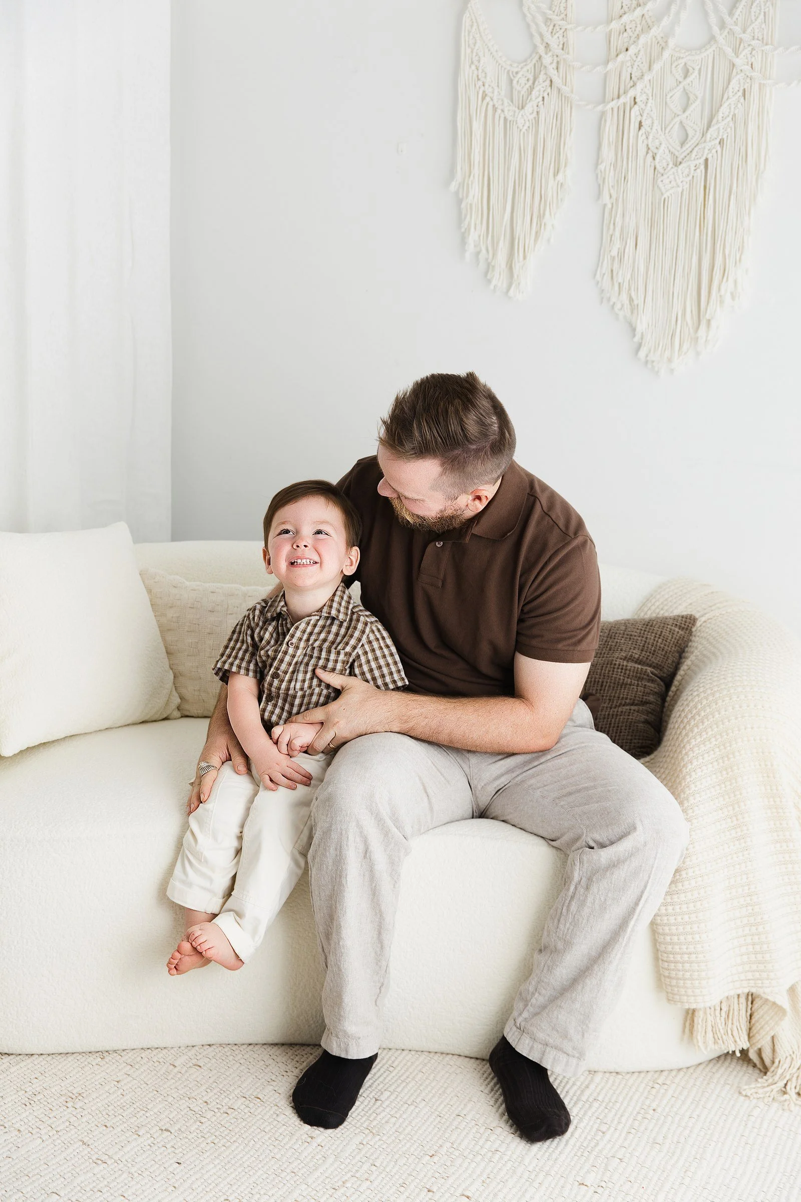 A smiling toddler in a checked shirt sitting beside his father on the studio sofa.