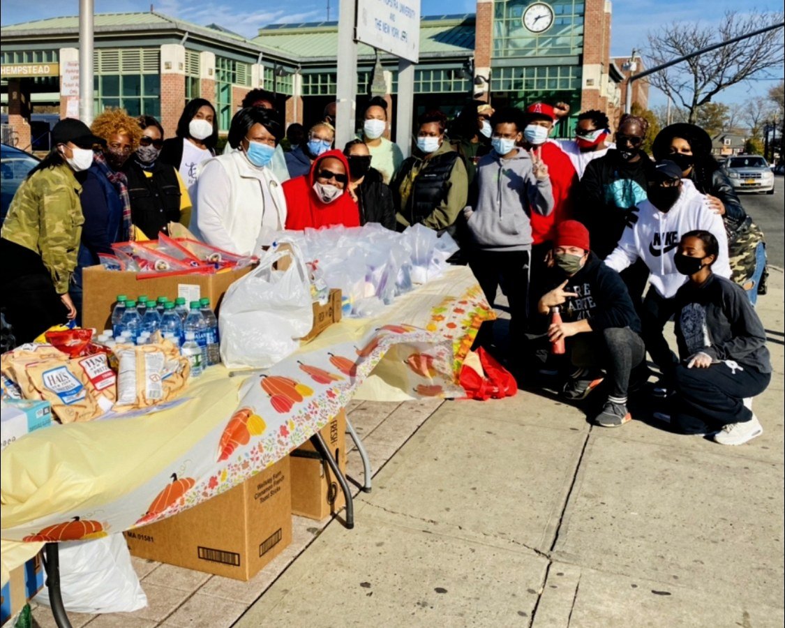 LFT members gather behind a sidewalk table of food, water, and care bags during outreach outside the Hempstead train station.