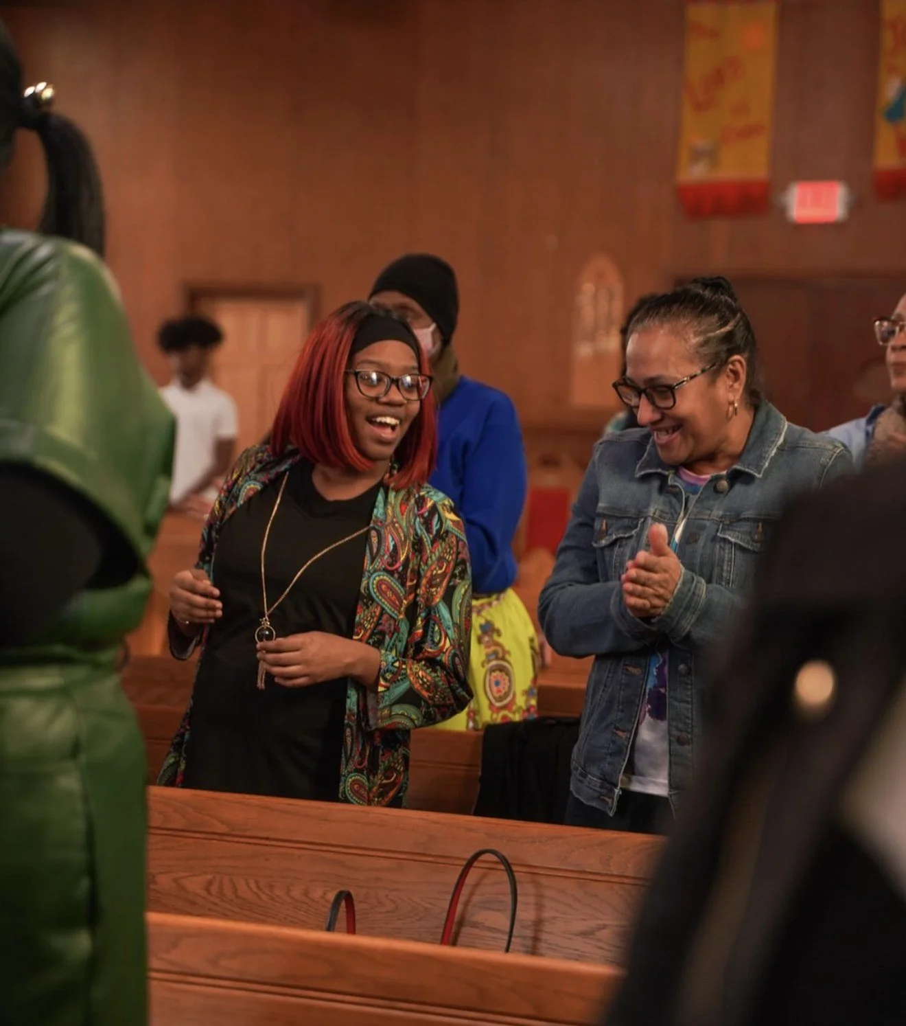 Two women smile and clap together in the pews during worship at LFT, with other congregants around them