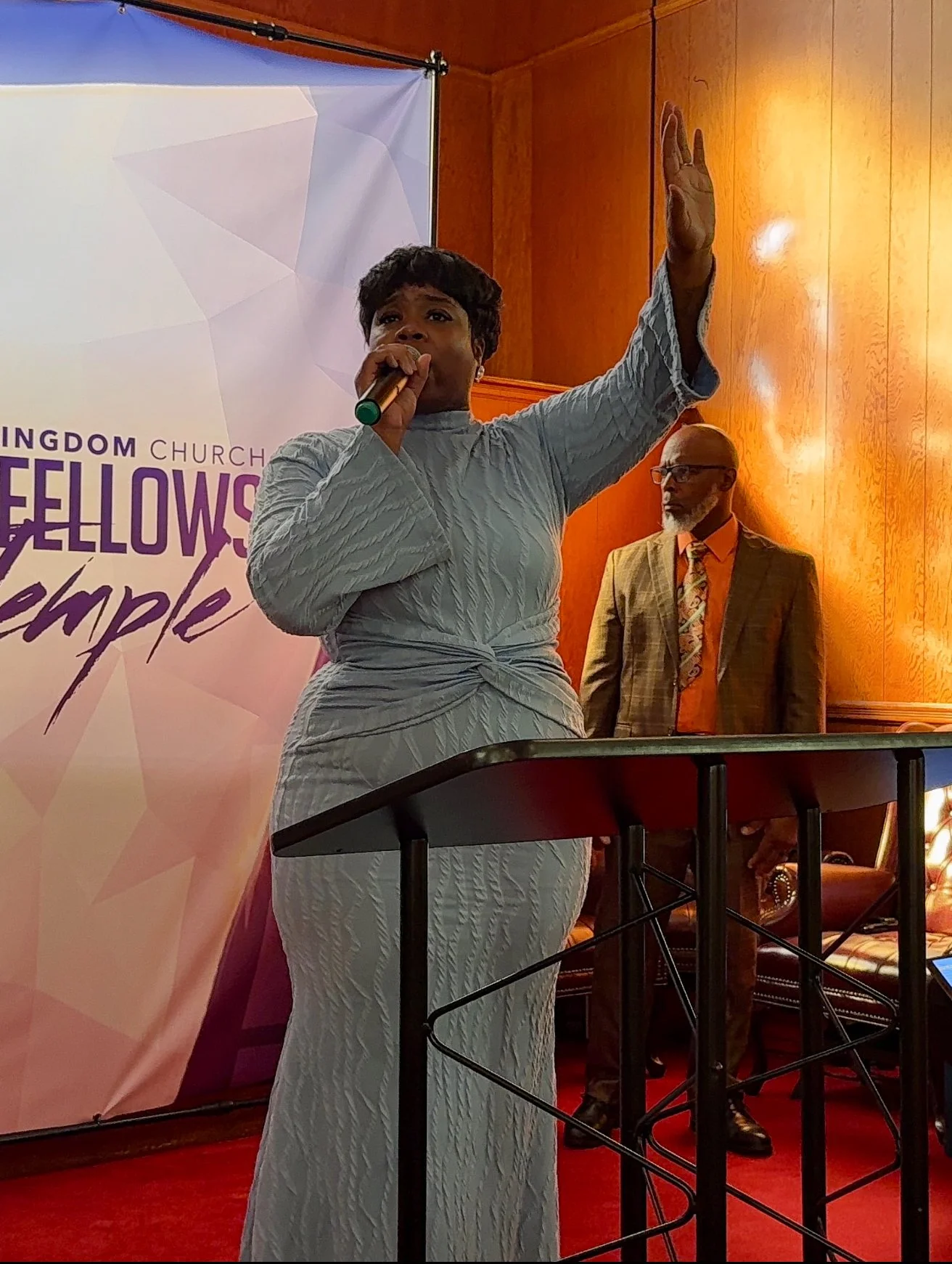 Pastor Olivia Simpkins speaks into a microphone with one hand raised, standing before The Kingdom Church | Love Fellowship Temple banner