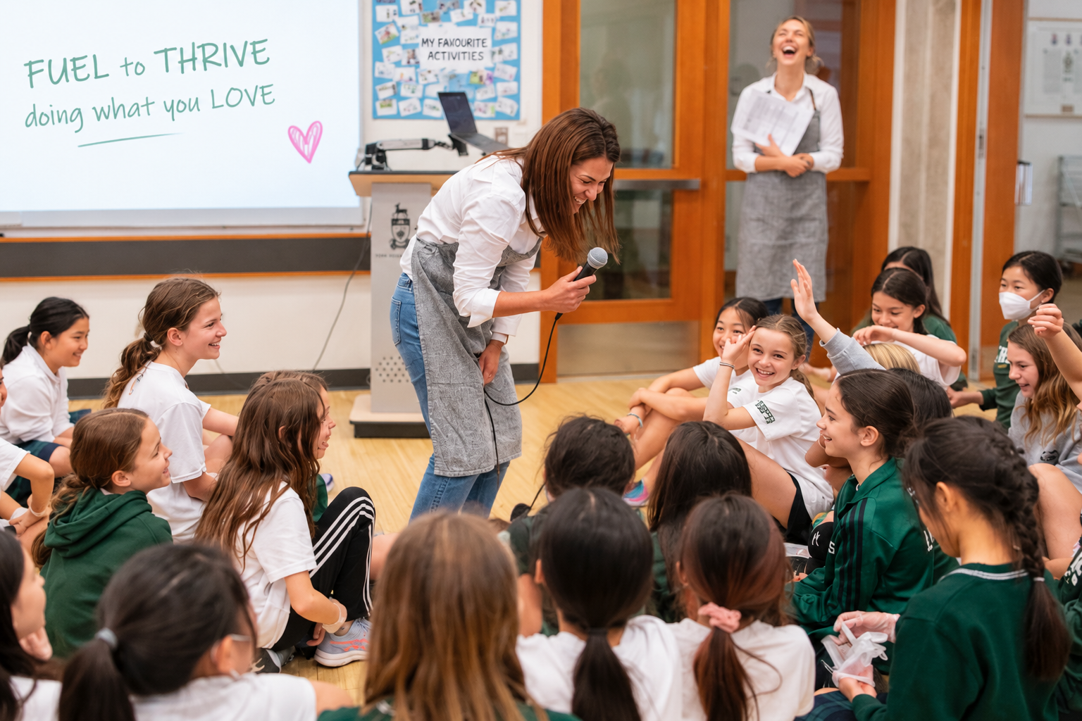 A nourishED facilitator leans in with a microphone, laughing with a circle of joyful students seated on the floor during a Fuel to Thrive workshop, with a second presenter laughing in the background
