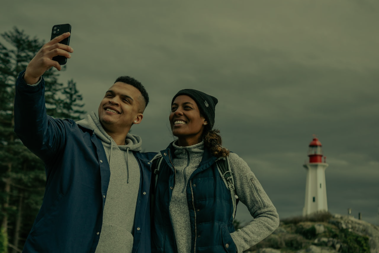 A young man and woman in their mid-twenties dressed in casual outdoor clothing take a selfie on rocks with a lighthouse in the background.