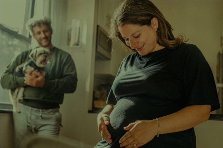 “Pregnant woman stands in her kitchen holding her stomach looking down smiling. In the background, a man standing by the kitchen window smiles at the woman while holding a small dog.