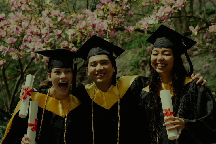 A young male, and two young females stand together with arms around each other and holding their diplomas dressed in graduation robes.