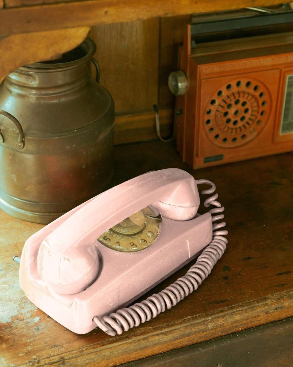 A pink rotary telephone on a wooden shelf, beside an old copper churn and an orange radio