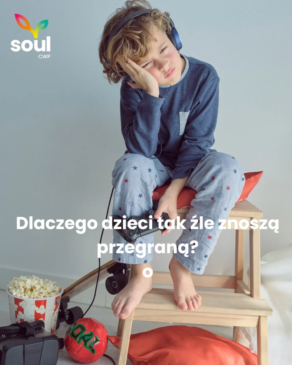 Smutny chłopiec w piżamie ze słuchawkami i padem do gry siedzi na drewnianym stołku, obok popcorn i piłka, z pytaniem: Dlaczego dzieci tak źle znoszą przegraną?