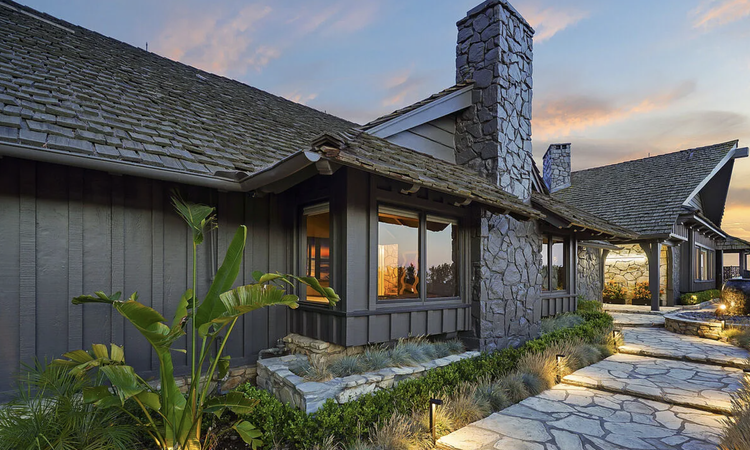 Dark board-and-batten ranch home with stone chimney at twilight