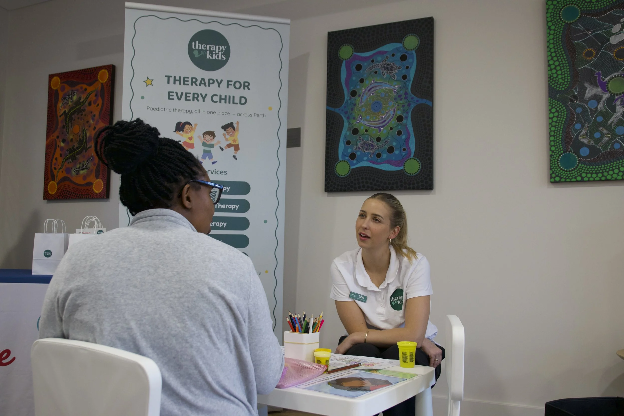 A Therapy for Kids Perth clinician sitting at a small table talking with a parent, with the Therapy for Kids Perth banner behind them.