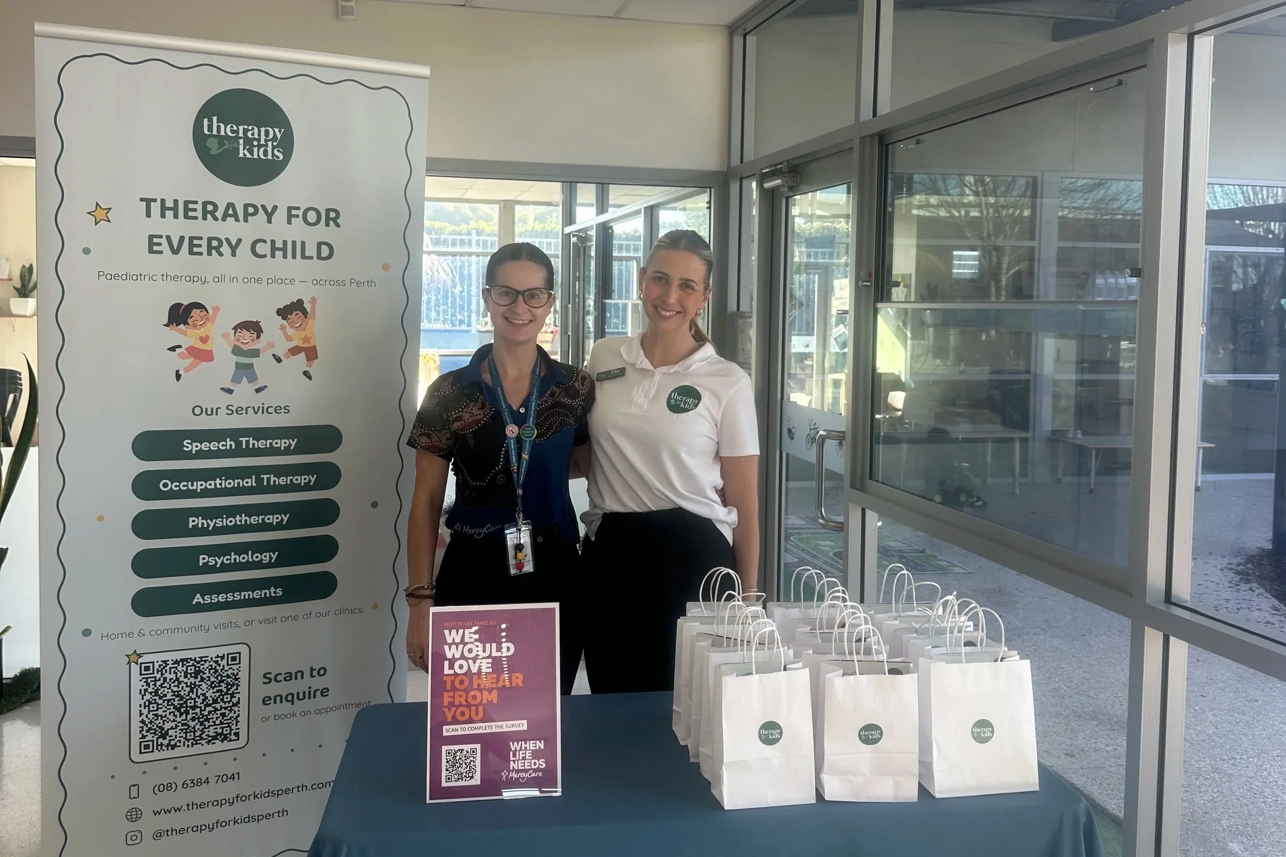A Therapy for Kids Perth clinician and a MercyCare Early Learning staff member standing together at an information stand at a MercyCare centre.