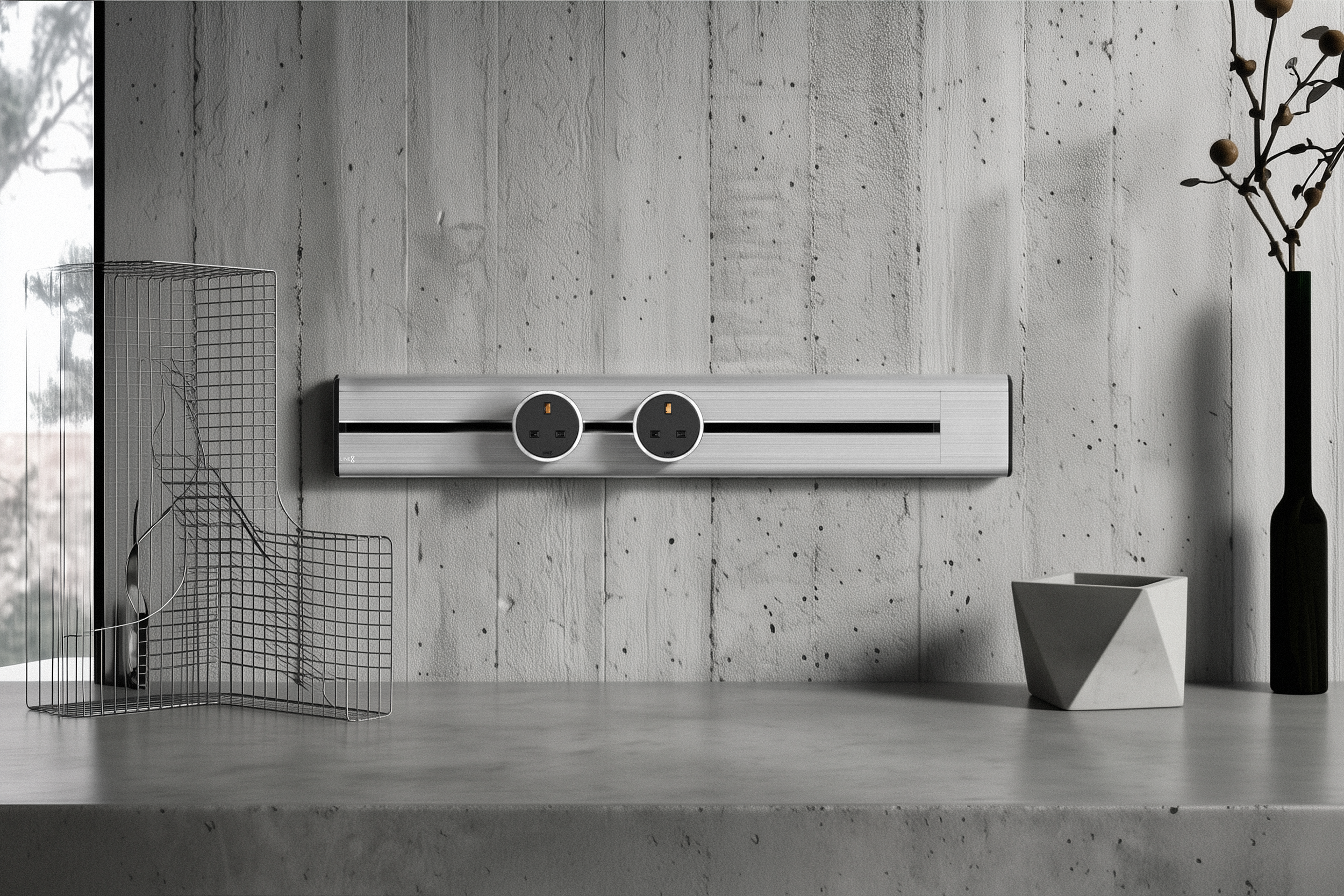 A detailed, architecturally-inspired close-up of the Line8 Model SL power track, finished in premium Silver Hairline brushed aluminum. The track is mounted horizontally on a textured industrial-chic concrete wall with formwork lines. The track body features two circular black power socket modules with central brass and copper contacts, set within a black channel. The scene is curated on a matching concrete countertop with sophisticated, minimalist objects: a complex geometric wire-mesh sculpture to the left, and to the right, a black vase with a dried branch next to a geometric-faceted pot. Natural light streams from a window on the far left, creating a high-end, bespoke interior aesthetic.