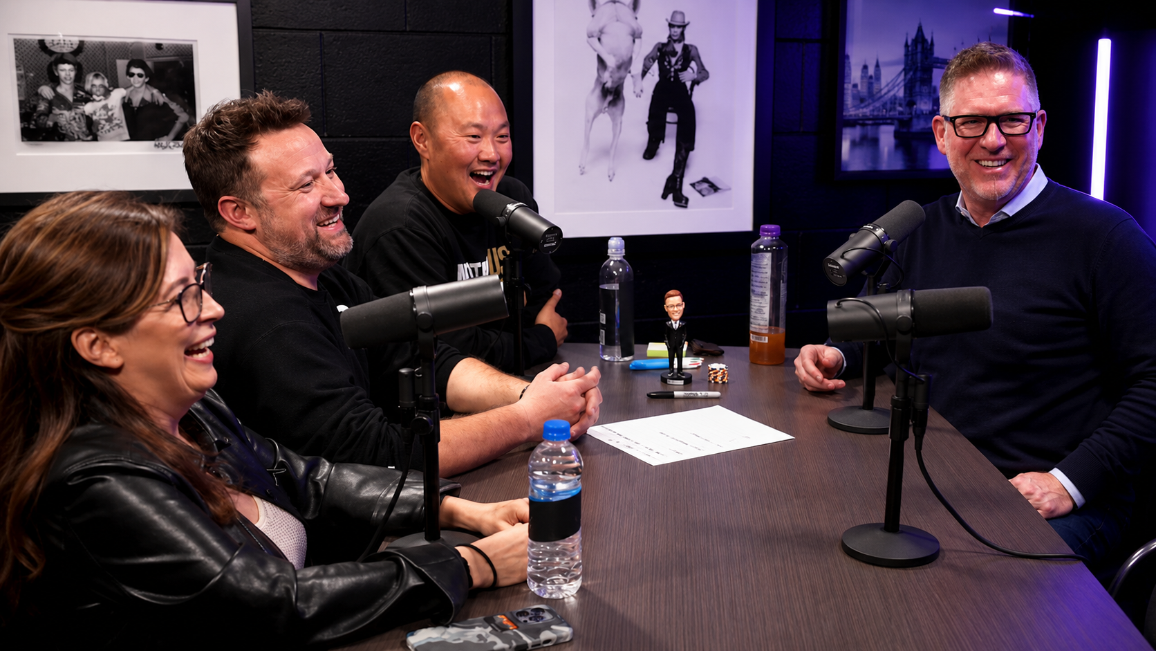Four people sitting at a table with microphones, laughing during a podcast recording.