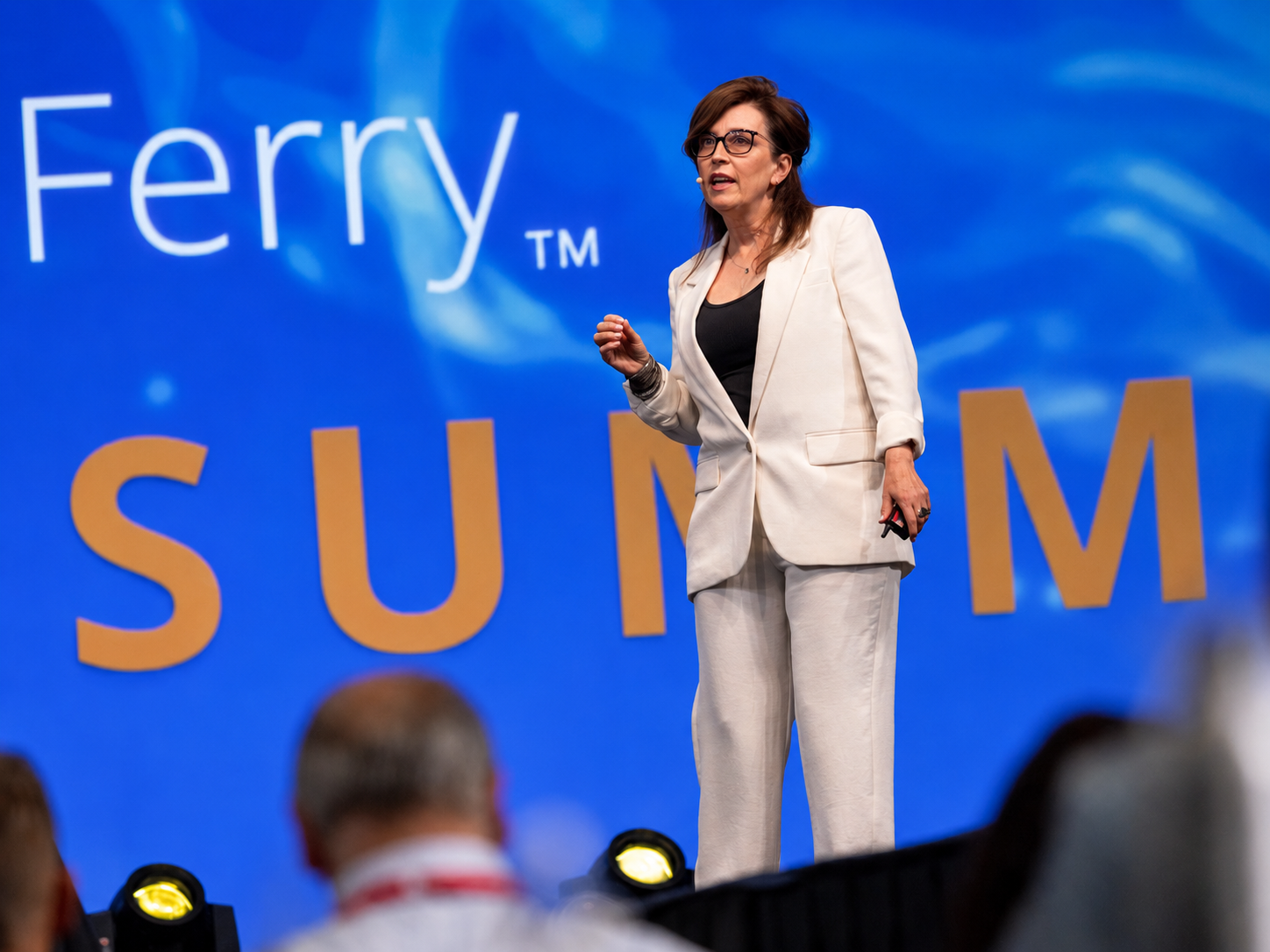 A woman in a white suit speaking on stage at a conference with a large blue background that says Tom Ferry SUMMIT in multicolored letters.