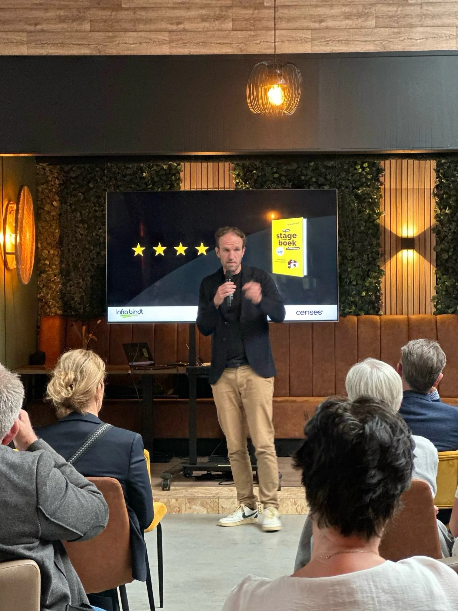 Maarten Brand spreekt voor een zaal, met Het Kleine Stageboek op het scherm achter hem