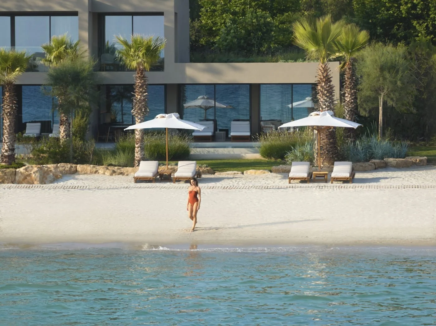 A woman walking along the water's edge in front of the Sani Asterias residences, with sun loungers and white parasols