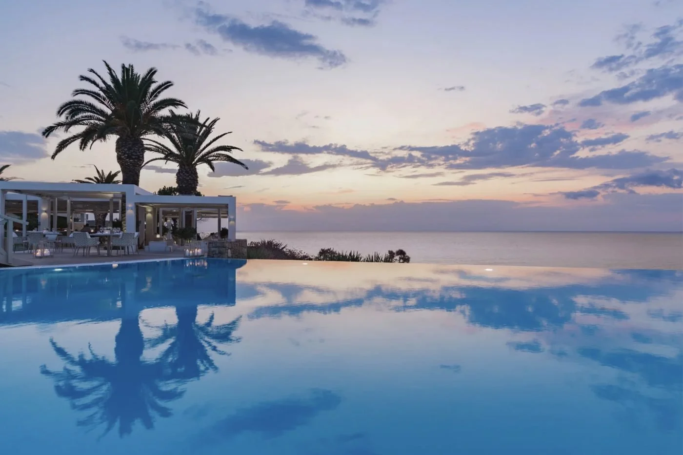 A candlelit restaurant terrace beside the infinity pool at Sani Club at sunset, with palm trees and the sea beyond