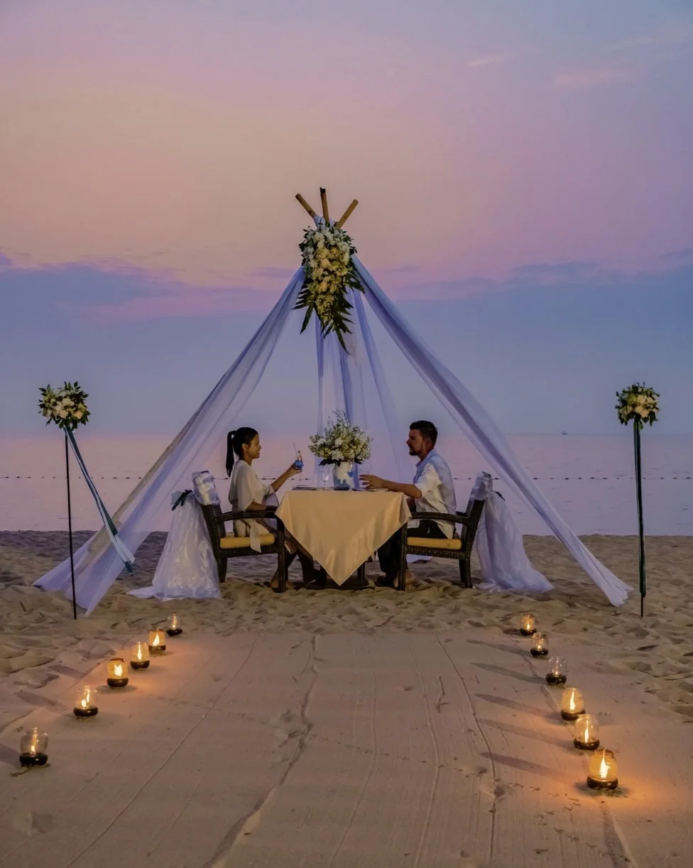 A candlelit table for two under a draped canopy on the beach at dusk
