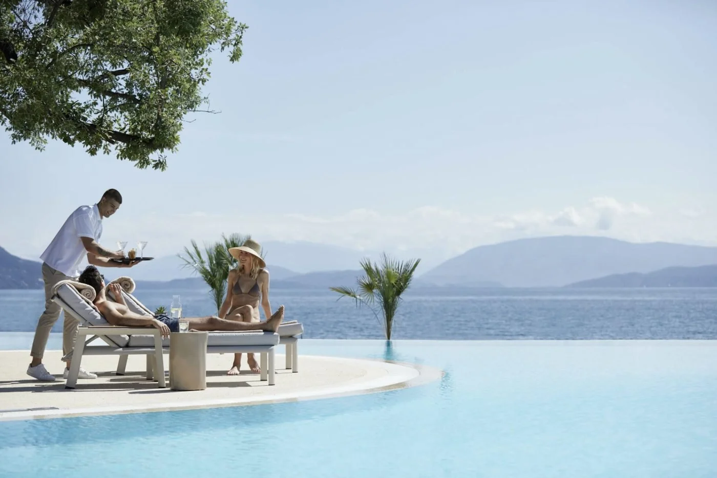 A waiter bringing drinks to guests on sun loungers beside an infinity pool overlooking the sea
