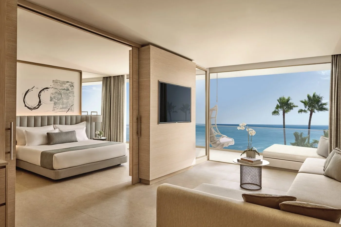 A Deluxe One Bedroom Suite Sea Front View with a hanging chair on the balcony and palm trees against the open sea