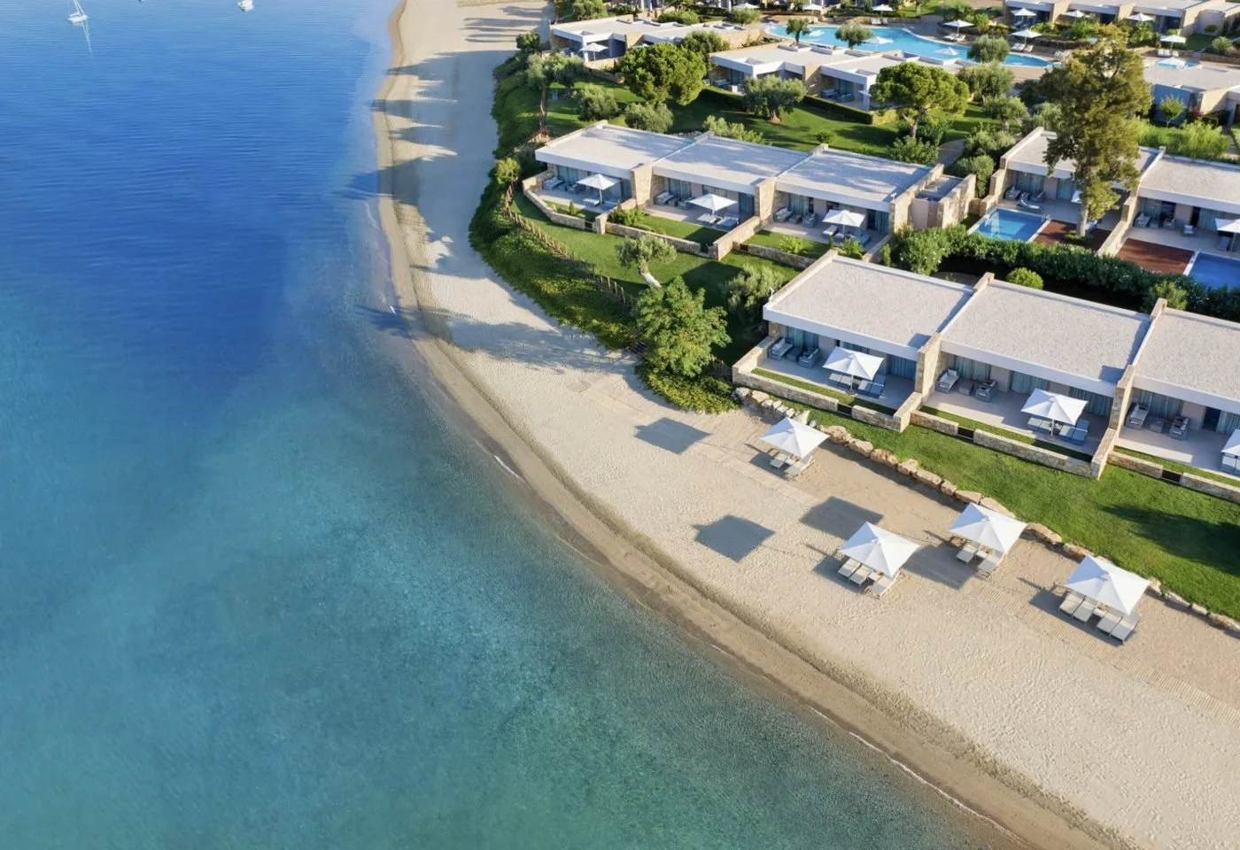 An aerial view of the beachfront Deluxe Collection suites at Ikos Olivia, with white parasols on the sand and the sea alongside