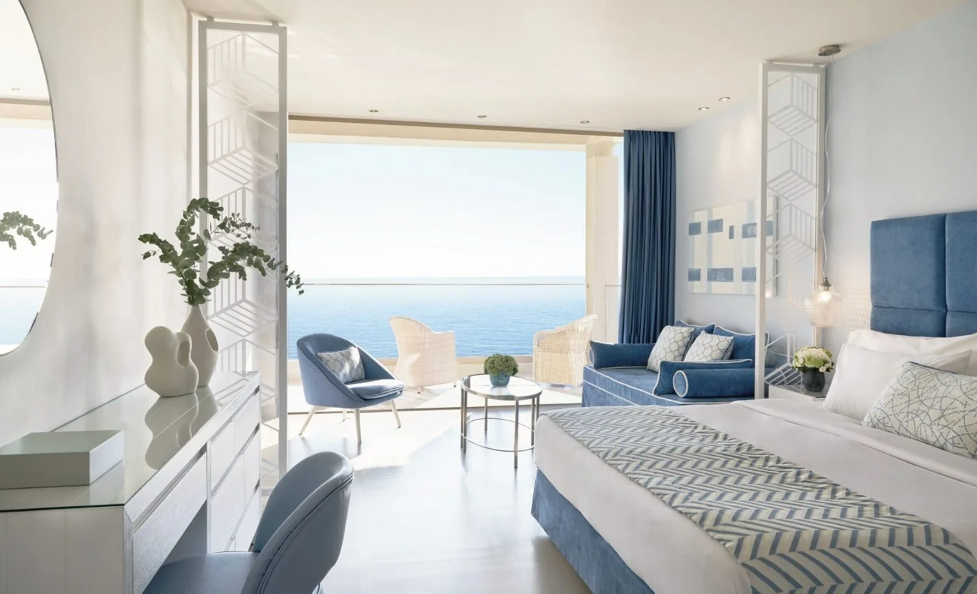 A Panorama Junior Suite at Ikos Oceania with a sitting area and wide windows onto a balcony over the sea