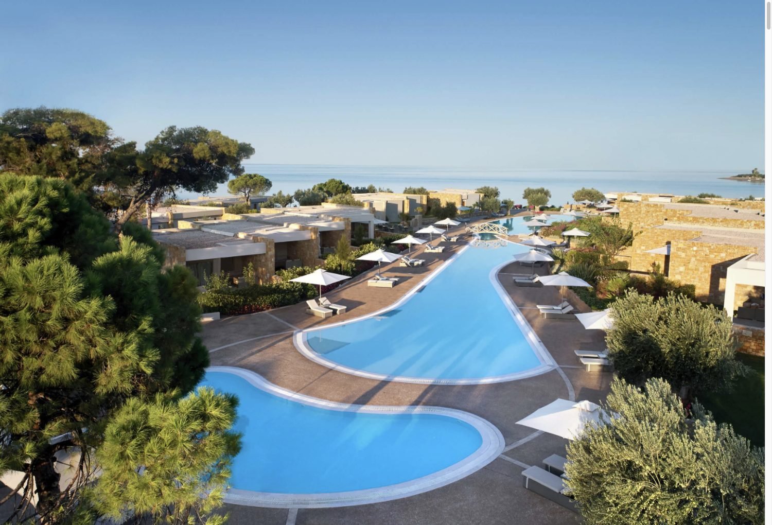 Lagoon pools winding between pine trees and stone buildings at Ikos Olivia in Halkidiki