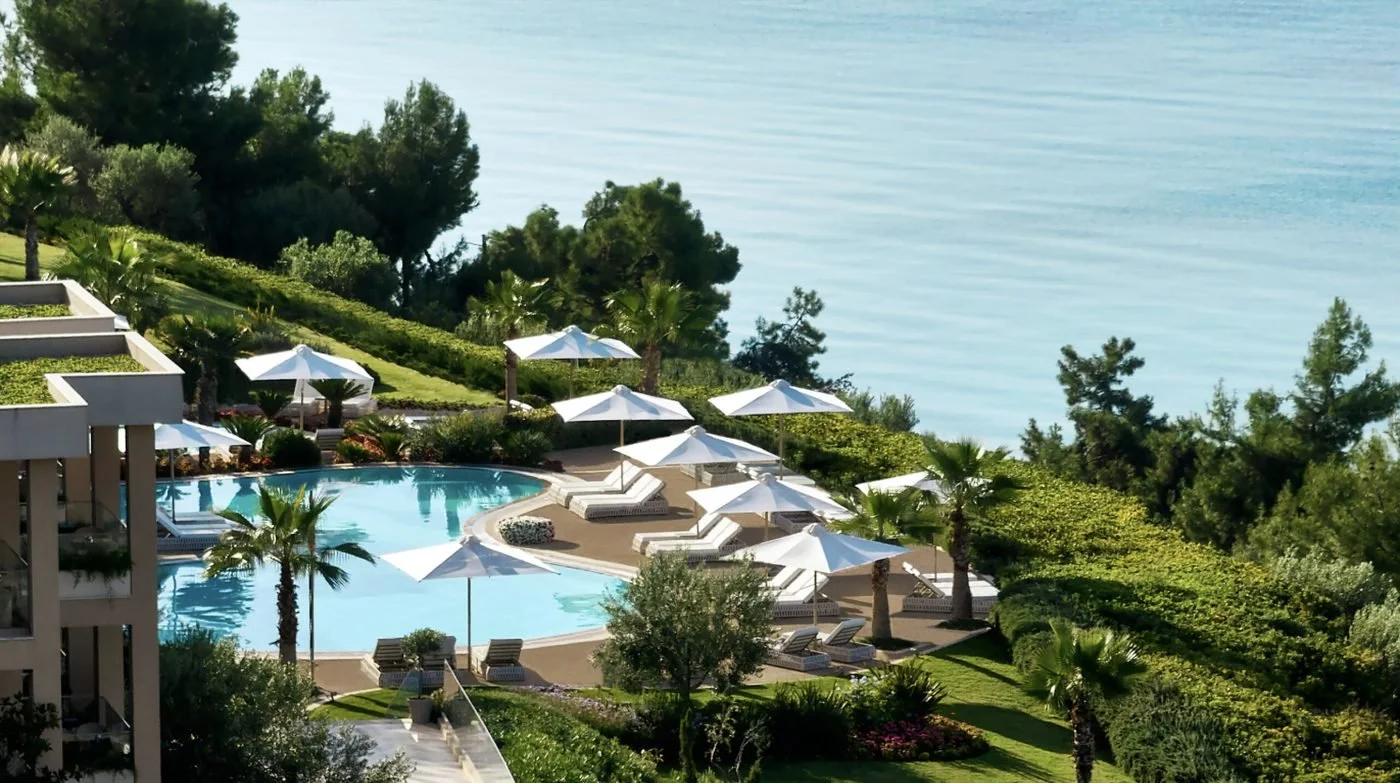 A pool surrounded by palms, parasols and gardens on the hillside at Ikos Oceania, looking out to sea