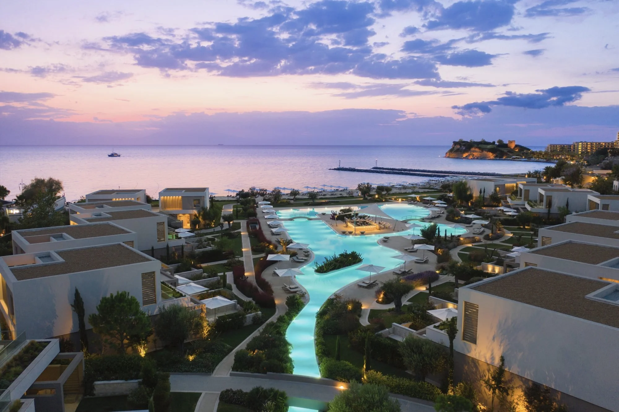 Sani Dunes at dusk from above: low-rise suites around the lit lagoon pool, with the beach, a wooded headland and the sea beyond