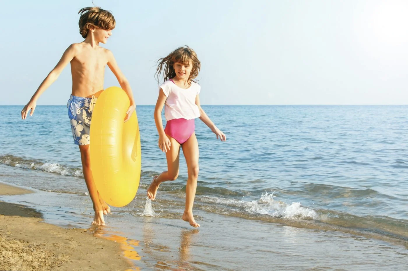 Two children running through the shallows on a sunny beach, one carrying a yellow rubber ring