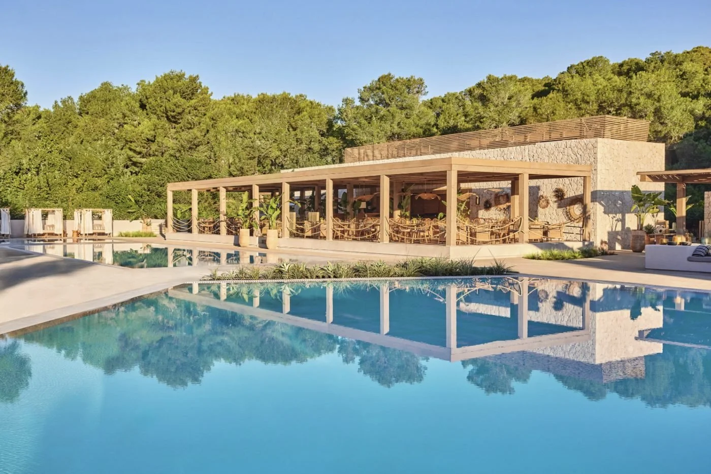 An open-sided restaurant pavilion reflected in a still pool, backed by pine trees