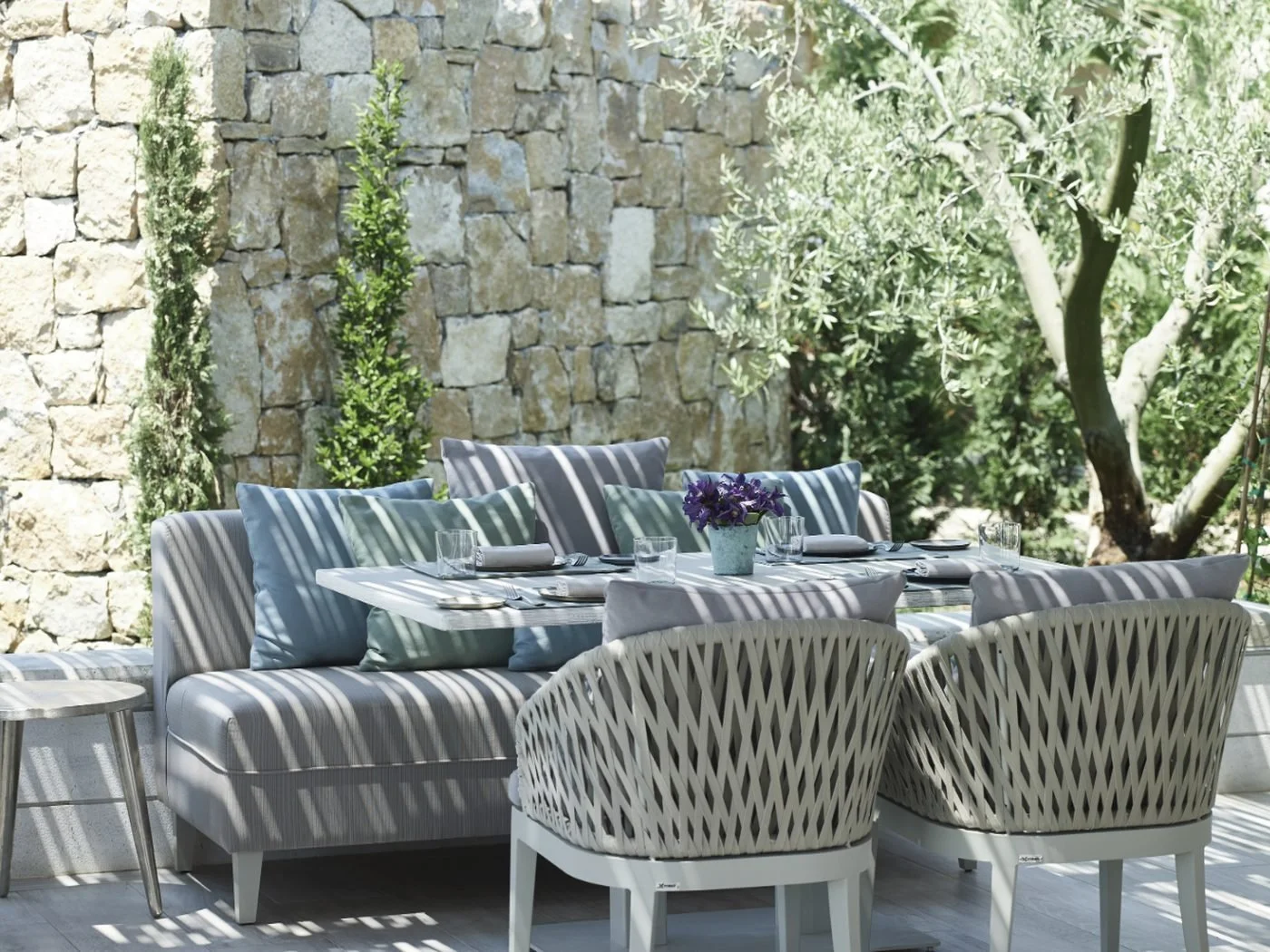 Cushioned sofas and woven chairs around a table at Beach House, beneath an olive tree and a stone wall