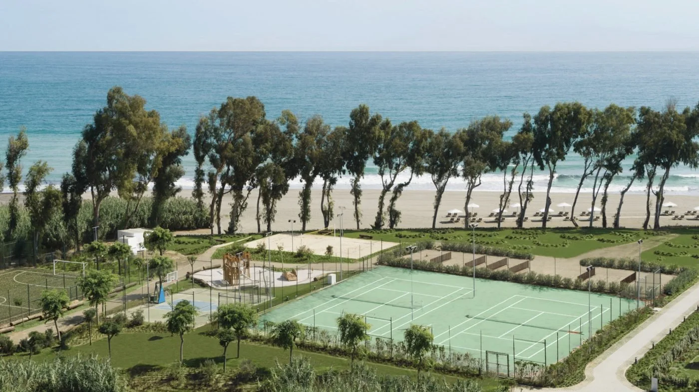 Tennis courts, a beach volleyball court and a playground in the gardens, with a line of trees and the beach beyond