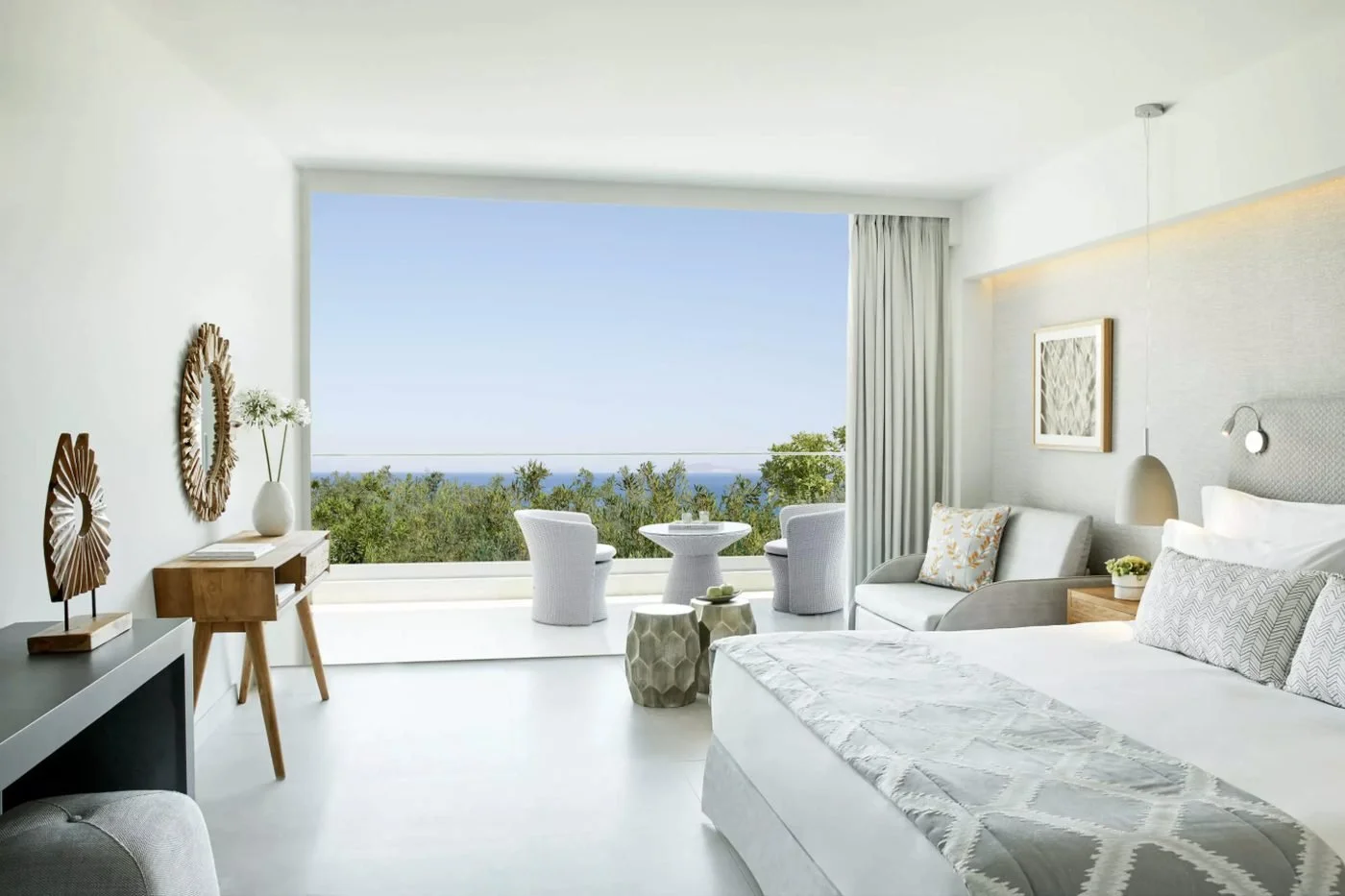 A Double Room at Ikos Aria with a wide picture window opening onto a furnished balcony above green treetops and the sea