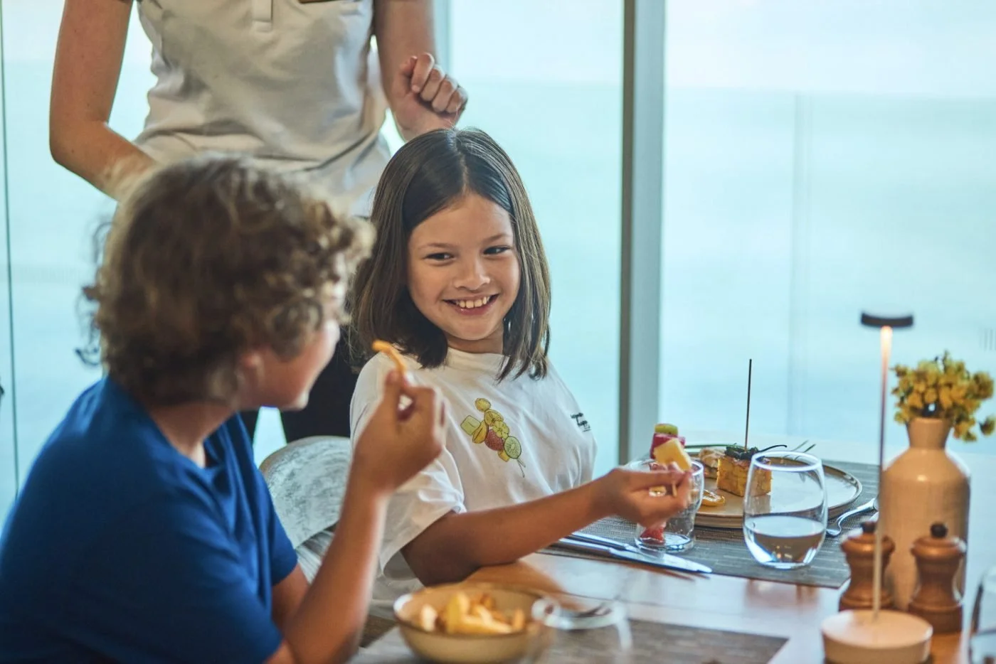 A smiling girl and a boy sharing a snack at a table by a window overlooking the sea