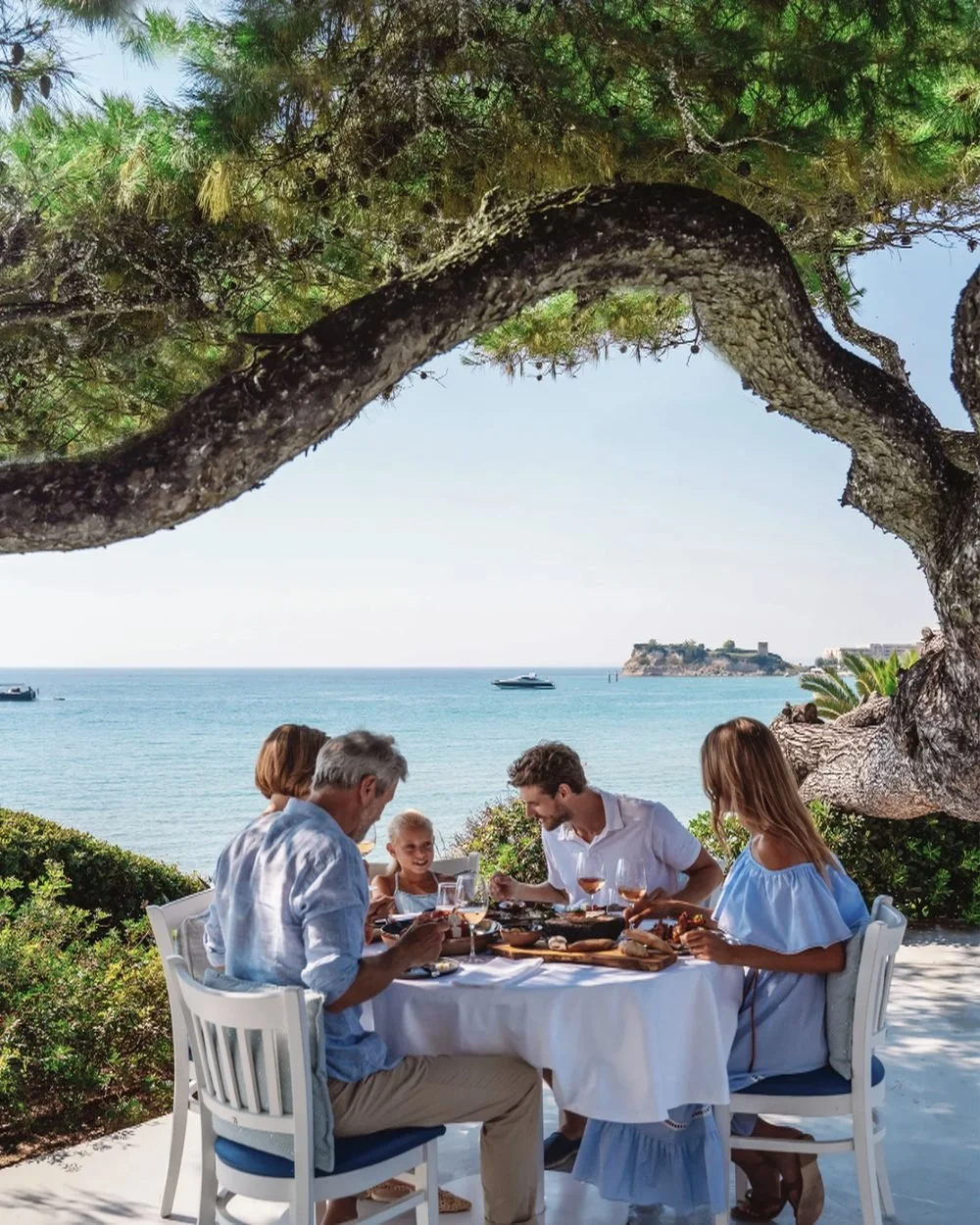 A multi-generational family sharing lunch under a pine tree overlooking the sea at Sani Club