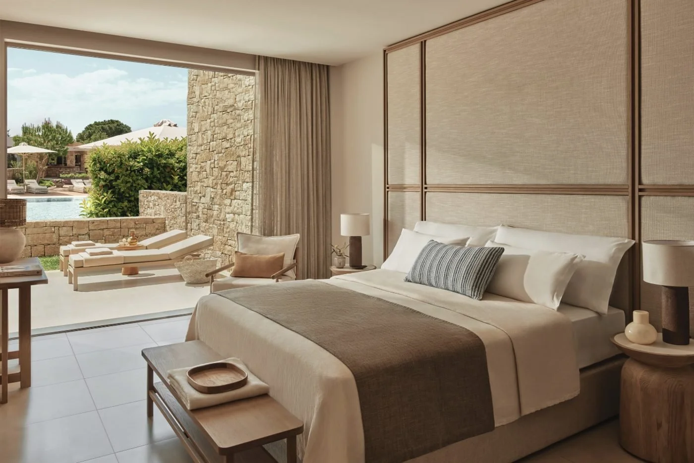 The bedroom of a Deluxe One Bedroom Bungalow Suite at Ikos Olivia opening onto a garden terrace, with a pool beyond