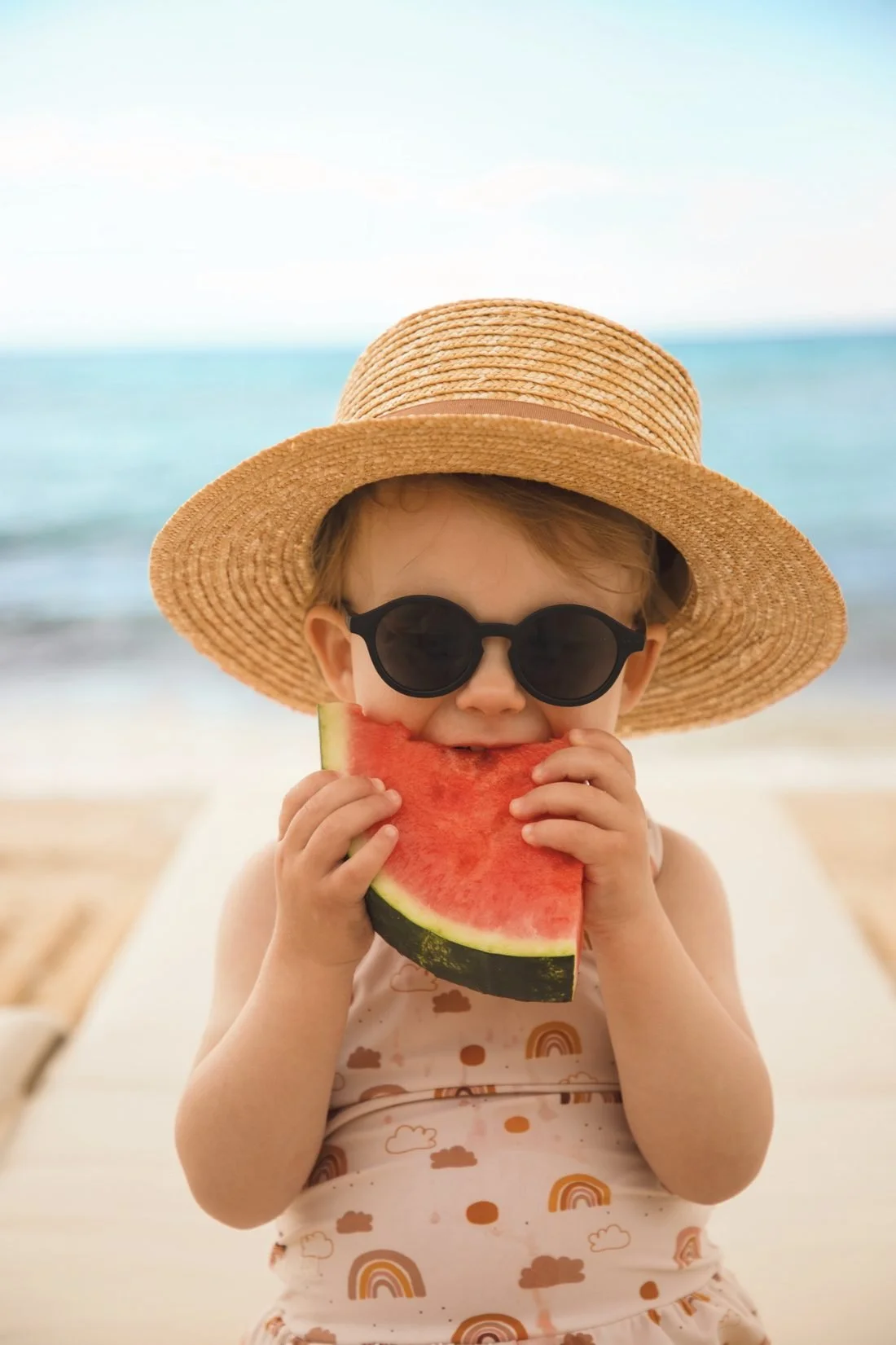 A toddler in a straw hat and sunglasses eating a slice of watermelon by the sea