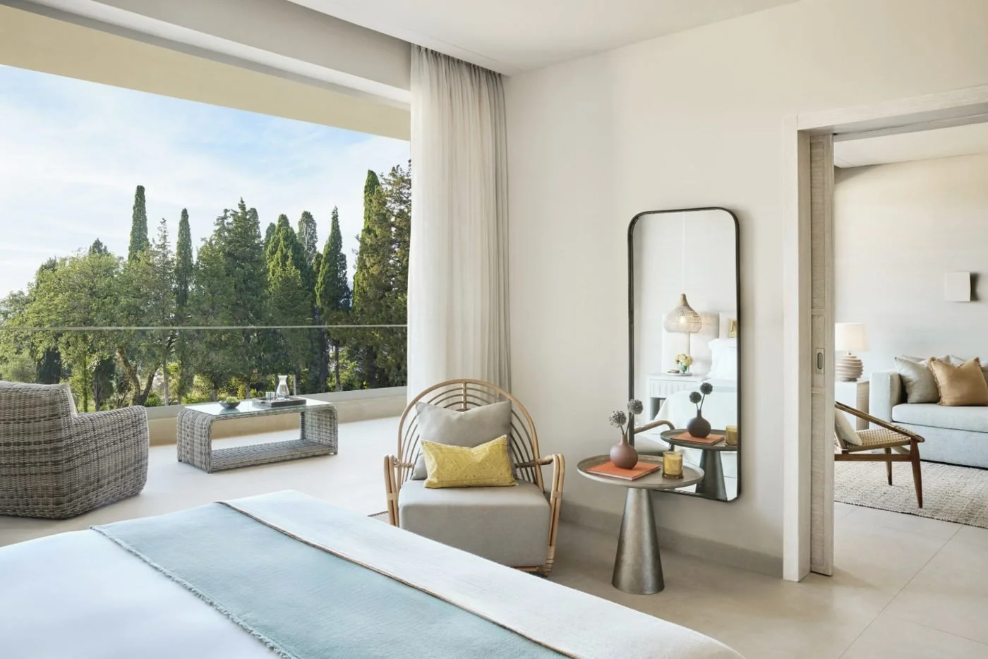 The bedroom of a Deluxe One Bedroom Bungalow Suite opening onto a balcony framed by cypress trees, with the living room beyond