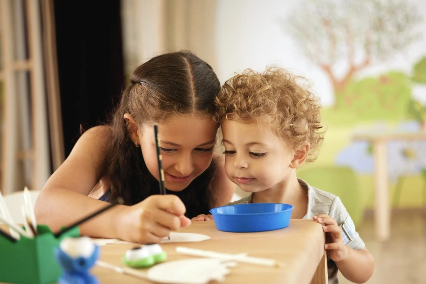 A girl and a little boy with curly hair painting together at a craft table